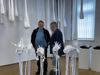 Zwei Männer stehen in einer Galerie und betrachten Kunstwerke. Um sie herum hängen weiße Skulpturen und weitere Objekte.