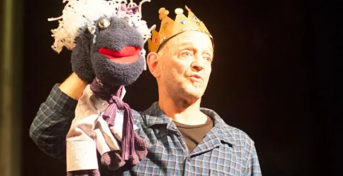 Ein Schauspieler präsentiert eine Handpuppe, die eine lustige Figur mit rotem Mund und weißen Haaren darstellt. Er trägt eine Krone und steht auf einer Bühne.