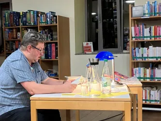 Ein Mann sitzt an einem Tisch in einer Bibliothek und liest etwas. Auf dem Tisch stehen Wasserflaschen und eine Lampe.