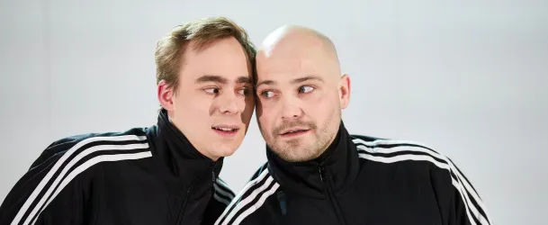 Zwei Männer in sportlichen Jacken stehen nah beieinander. Sie sehen einander an und wirken neugierig.