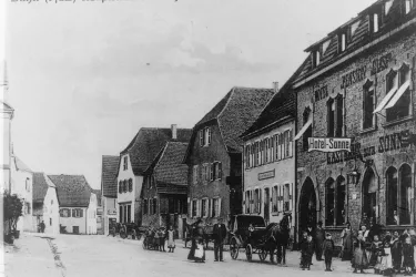 Eine historische Straßenszene mit mehreren Gebäuden. Menschen und Pferdewagen sind ebenfalls zu sehen.