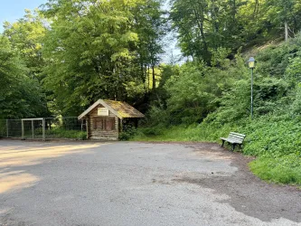 Eine kleine, holzige Hütte steht in einem grünen Waldgebiet. Daneben gibt es eine Parkbank und eine Straßenlaterne.