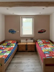 Ein helles Kinderzimmer mit zwei Betten und bunten Bettwäsche. An der Wand hängen Bilder von Autos.