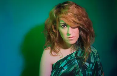 Eine Frau mit lockigem, braunem Haar und einem grünen Outfit schaut direkt in die Kamera. Der Hintergrund ist in einem sanften Grünton gehalten.