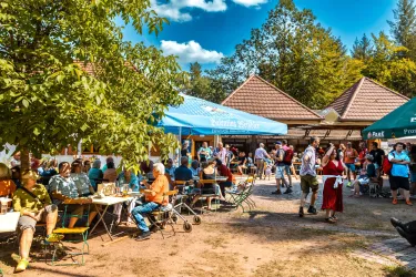 Ein lebhaftes Fest mit vielen Menschen, die an Tischen sitzen und essen. Im Hintergrund sind Zelte und Bäume zu sehen, die eine einladende Atmosphäre schaffen.