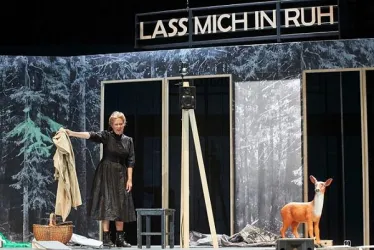 Eine Schauspielerin steht auf einer Bühne und hält einen Mantel in der Hand. Neben ihr steht ein kleines Reh und im Hintergrund sieht man eine Waldlandschaft mit dem Schriftzug „LASS MICH IN RUH“.