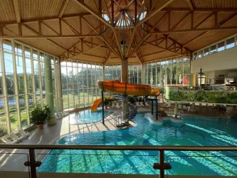 Ein Innenbereich mit einem großen Schwimmbecken und einer Wasserrutsche. Große Fenster lassen viel Tageslicht herein und bieten einen Blick auf die Außenlandschaft.