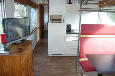 Eine moderne, helle Wohnküche mit einem Fernseher, einem kleinen Tisch und einer Kochnische. Die Einrichtung ist einfach und funktional, mit Holz- und Metallakzenten.
