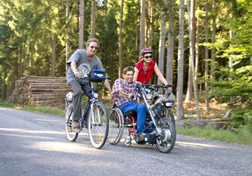 Drei Personen fahren mit Fahrrädern und einem speziellen Handbike auf einem Waldweg. Die Umgebung ist naturnah, mit hohen Bäumen im Hintergrund.