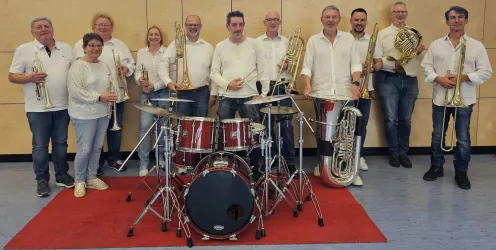 Eine Gruppe von Musikern steht zusammen mit Instrumenten in einem Probenraum. Im Vordergrund befindet sich ein Schlagzeug.