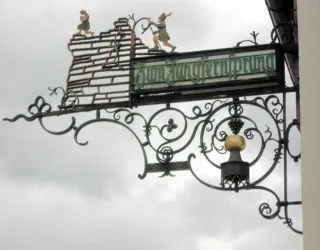 Ein kunstvoller Metallladen mit einer Schilderung über einem Gebäude. Die Darstellung zeigt zwei Figuren, die auf einer Mauer stehen.