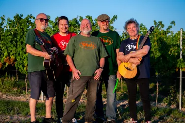 Eine Band steht in einem Weinberg, umgeben von Traubenreben. Sie halten Musikinstrumente und lächeln in die Kamera.