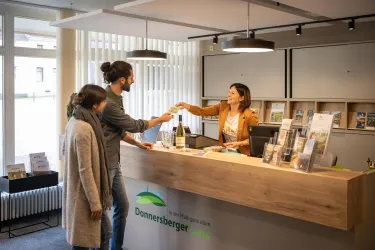 Eine Empfangstheke mit einer freundlichen Mitarbeiterin, die einem Kunden eine Flasche Sekt überreicht. Im Hintergrund stehen Informationsbroschüren und es sind zwei weitere Personen anwesend.