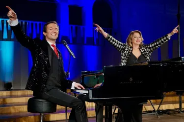 Ein Duo von Sängern auf der Bühne, die mit Begeisterung ihre Arme heben. Sie stehen an einem Klavier und präsentieren eine gesellige Aufführung.