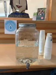 Ein großer Glaskrug mit klarem Wasser steht auf einem Tisch. Neben dem Krug befinden sich Plastikbecher und ein Schild mit der Aufschrift "Refill Station".