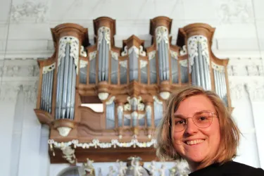 Eine Person lächelt vor einer großen Orgel. Die Orgel ist kunstvoll gestaltet und hat viele Pfeifen.