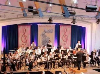 Ein Blasorchester spielt auf einer Bühne mit buntem Licht. Im Hintergrund sind Musikinstrumente und Notenblätter sichtbar.