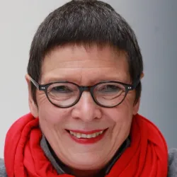 Eine lächelnde Frau mit kurzen, grauen Haaren und einer roten Schal. Sie trägt eine Brille und hat einen freundlichen Ausdruck.