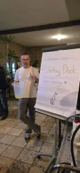 Ein Mann steht neben einem Flipchart mit der Aufschrift "Willkommen zum Walking Duck". Er hält einen Behälter in der Hand und ist in einem Raum mit Pflanzen.