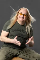 Ein lächelnder Mann mit langen, blonden Haaren sitzt auf einem Hocker. Er trägt eine grüne T-Shirt und auffällige gelbe Sonnenbrille.