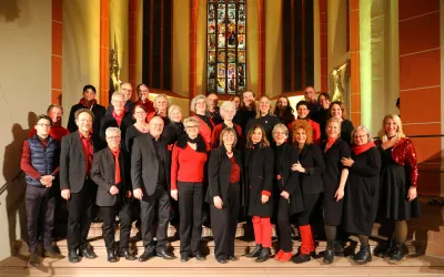 Eine Gruppe von Sängern steht auf einer Treppe in einer Kirche. Sie tragen schwarze Kleidung mit roten Akzenten und lächeln in die Kamera.