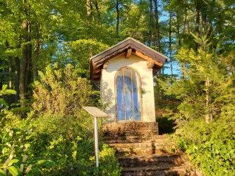 Ein kleiner Marienaltar umgeben von grünen Bäumen und Sträuchern. Die Sonneneinstrahlung verleiht der Szene eine warme Atmosphäre.