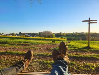 Zwei Paar Füße entspannen auf einer Bank mit Blick auf eine grüne Landschaft. Im Hintergrund sind sanfte Hügel und ein Wegweiser zu sehen.