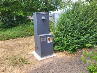 Ein grauer Trinkbrunnen steht auf einem hellen Platz im Freien. Umgeben ist er von Büschen und Gras.