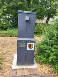 Ein grauer Trinkbrunnen steht auf einem Betonfundament. Umgeben von grünem Gras und Sträuchern in einem Wohngebiet.