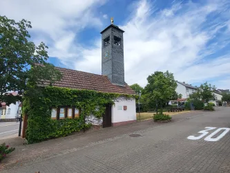 Ein kleines Gebäude mit einem Uhrturm steht an einer ruhigen Straße. Umgeben von Bäumen und einer freundlichen Nachbarschaft.