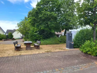 Ein ruhiger Platz mit einem Tisch und Bänken aus Holz sowie einem Trinkbrunnen, umgeben von Bäumen. Die Umgebung ist grün und einladend.