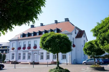 Ein schönes Gebäude mit einem roten Dach und weißen Wänden. Rundherum stehen gepflegte Bäume und ein Parkplatz ist sichtbar.