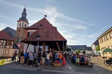Ein belebter Marktplatz mit vielen Menschen und einem historischen Gebäude. Im Hintergrund ist ein erhöhter Kirchturm zu sehen.