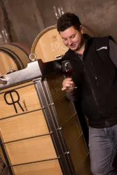 Ein Mann steht neben einem großen Weinfass und hält ein Glas Wein in der Hand. Er schaut lächelnd auf den Wein und befindet sich offensichtlich in einem Weinkeller.