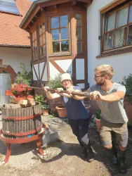 Zwei Personen drücken gemeinsam Äpfel in einer traditionellen Mostpresse. Im Hintergrund ist ein charmantes, altes Gebäude zu sehen.