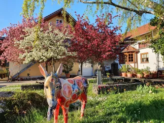 Ein buntes Esel-Bild steht auf einer Wiese vor einem schönen Haus. Im Hintergrund blühen pinke und weiße Bäume.