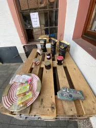 Ein Holztisch mit verschiedenen Lebensmitteln, darunter Snacks und Marmelade. Im Hintergrund sind Fenster und ein Schild zu sehen.