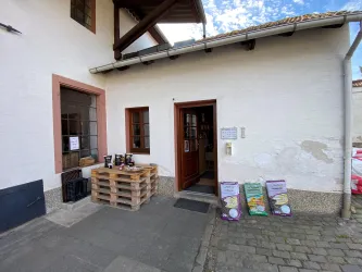 Ein Eingang zu einem Gebäude mit einem Holzregal aus Paletten und einigen Produkten davor. An der Wand befinden sich Schilder und das Wetter ist klar und sonnig.