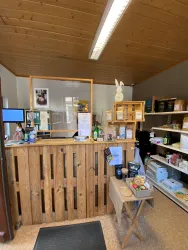 Ein Empfangsbereich mit einem Holzschalter und verschiedenen Regalen voller Produkte. An der Wand hängt ein Foto und es gibt einen Computer an der Theke.