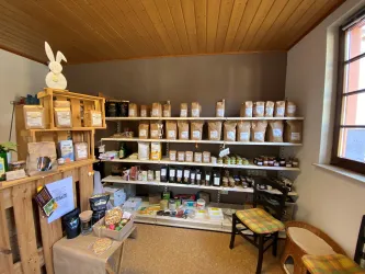 Ein gemütlicher Laden mit Regalen voller gehaltener Lebensmittel und Produkten. Im Vordergrund sind Holzkisten und ein Stuhl zu sehen.