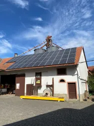 Ein modernes Gebäude mit Solarpanelen auf dem Dach. Der Himmel ist klar und blau, und die Umgebung wirkt ländlich.