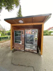 Ein Verkaufsautomat unter einem Holzüberdach. Neben dem Automaten befindet sich ein weiterer regionaler Verkaufsautomat.