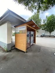 Ein Regiomat aus Holz steht an einem ruhigen Ort. Er bietet Produkte aus der Region an und ist von Bäumen umgeben.