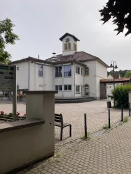 Ein modernes Gebäude mit einem Uhrenturm steht in einem gepflasterten Platz. Es sind einige Bäume und eine Parkbank in der Nähe zu sehen.