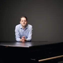 Ein Mann in einem blauen Hemd steht an einem Piano. Der Hintergrund ist schlicht und dunkel.