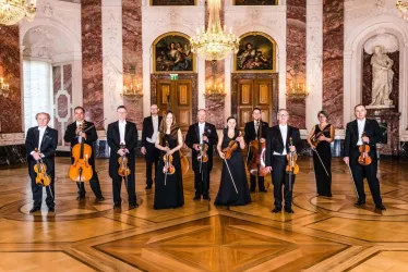 Eine Gruppe von Musikern steht in eleganter Kleidung in einem prächtigen Raum. Sie halten ihre Instrumente und strahlen eine festliche Atmosphäre aus.