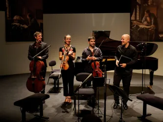 Ein Musikerensemble steht auf der Bühne und applaudiert nach einem Auftritt. Im Hintergrund sind Stühle und ein Klavier zu sehen.