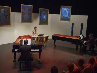 Ein Musikensemble spielt auf der Bühne, während ein Publikum zuschaut. Im Hintergrund hängen Bilder an der Wand und die Atmosphäre ist kreativ und künstlerisch.