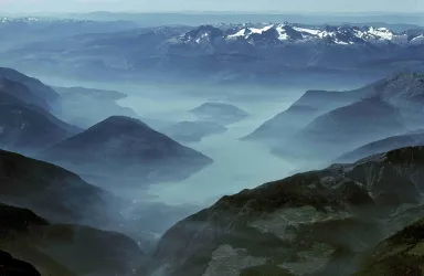 Eine beeindruckende Landschaft mit Berggipfeln und einem nebligen Tal. Der Blick zeigt ruhige Gewässer und eine fernere Bergkette.