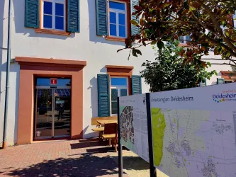 Ein Gebäude mit blauen Fensterläden und einer Eingangstür. Neben dem Gebäude steht eine Infotafel mit Karteninformationen über die Region Deidesheim.